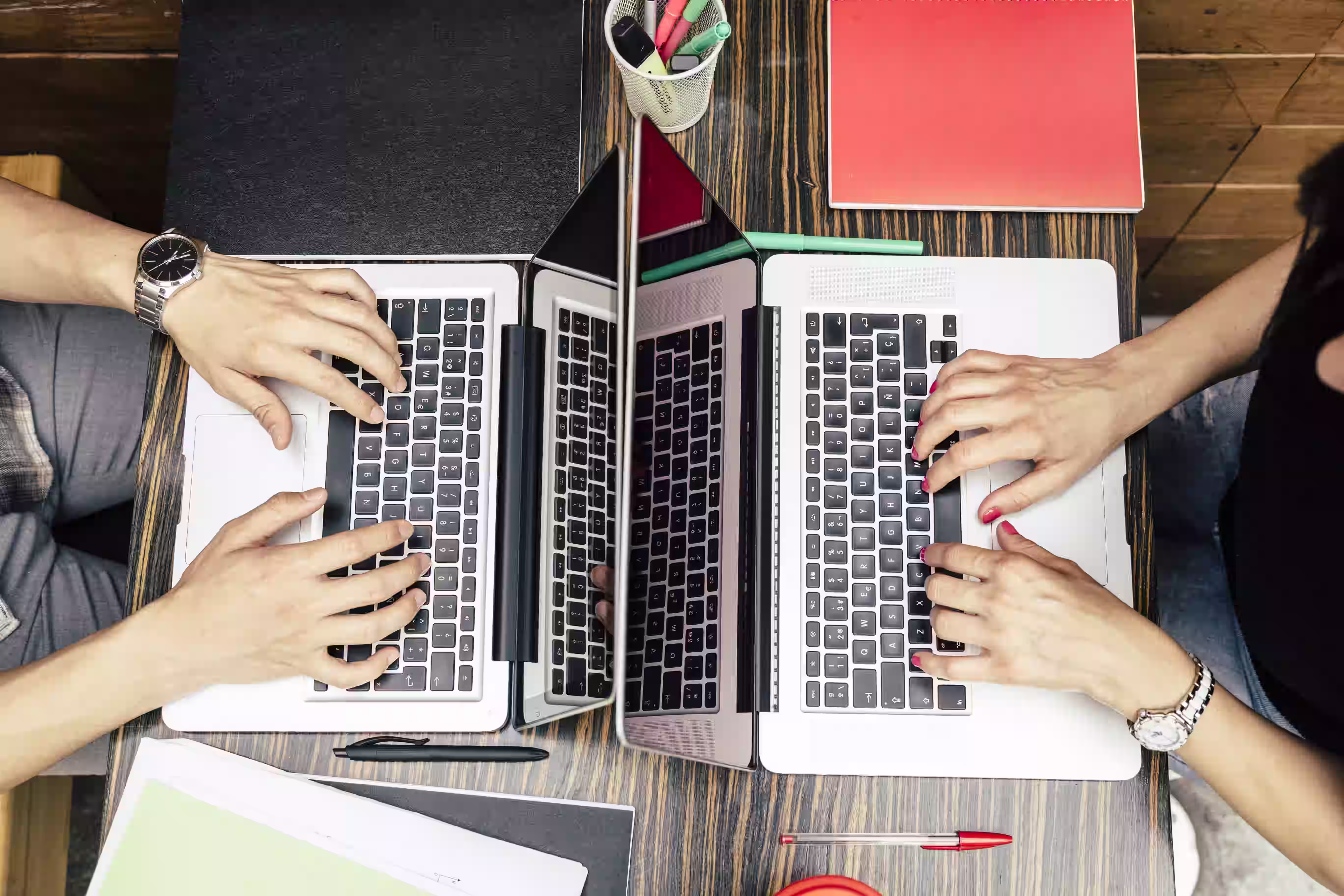 Overhead view of three researchers typing on laptops at a wooden table with stationery