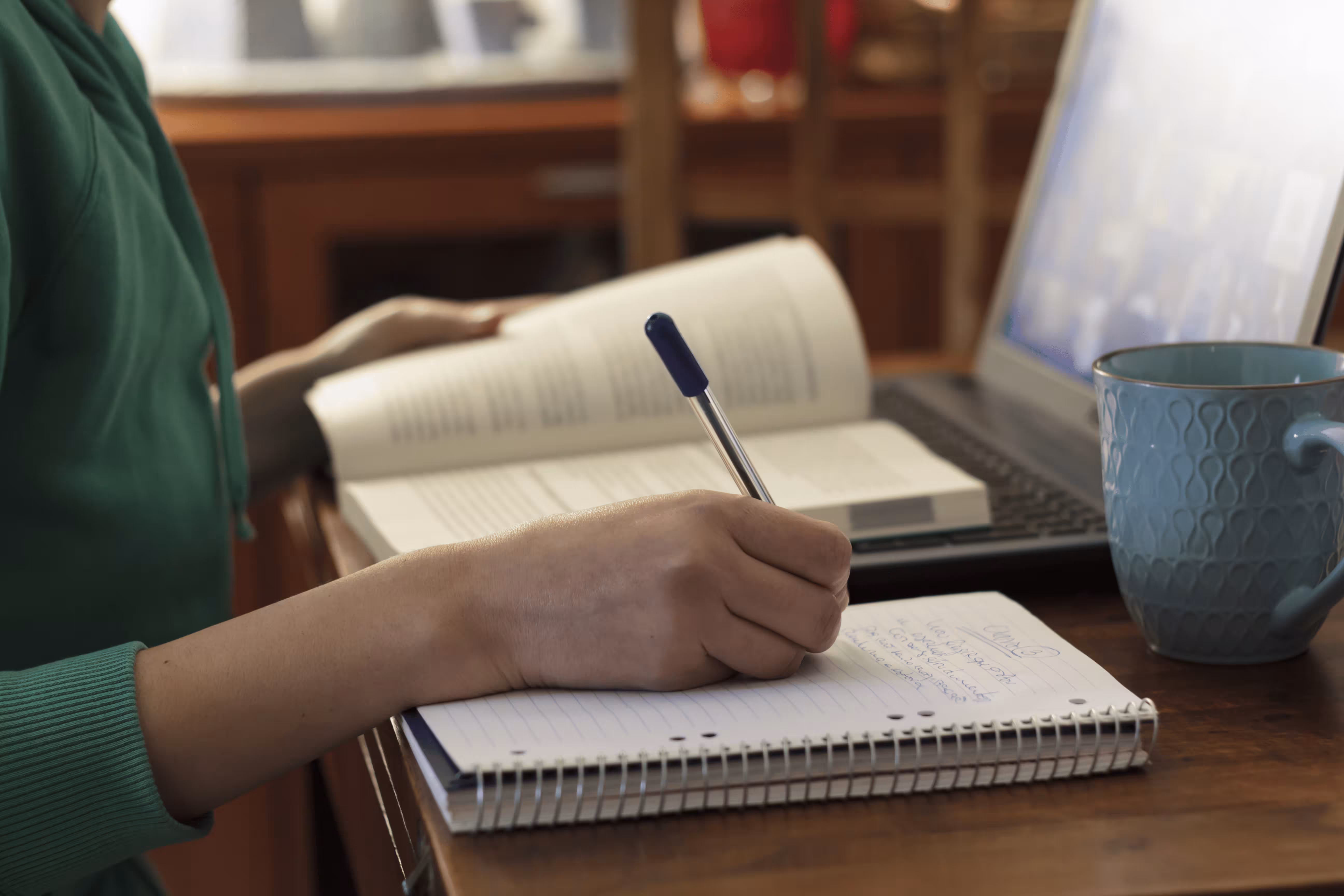 Person writing in spiral notebook with open book and laptop in background